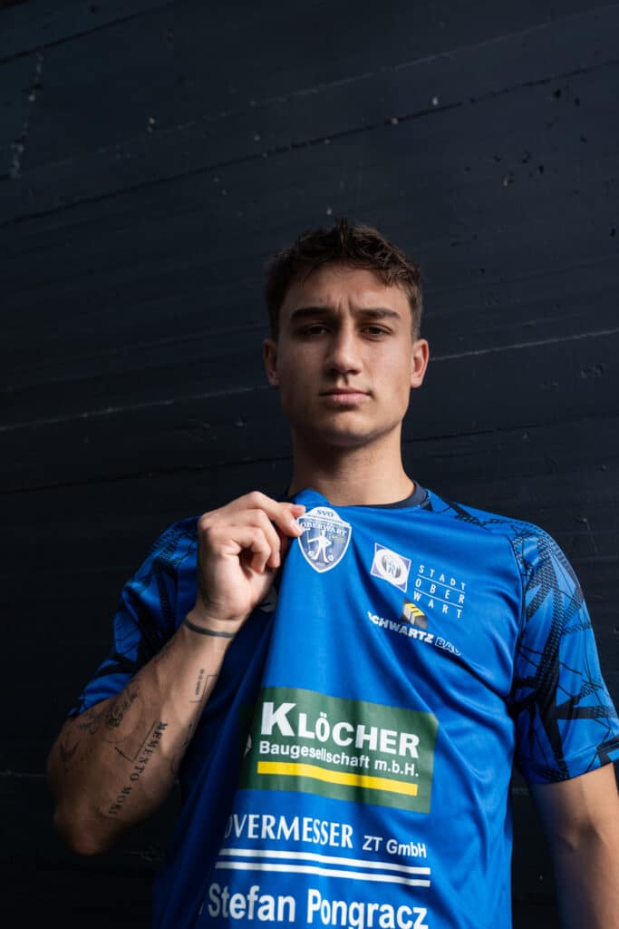 Junger Mann im blauen Fußballtrikot hält Vereinslogo vor dunklem Hintergrund in die Kamera.