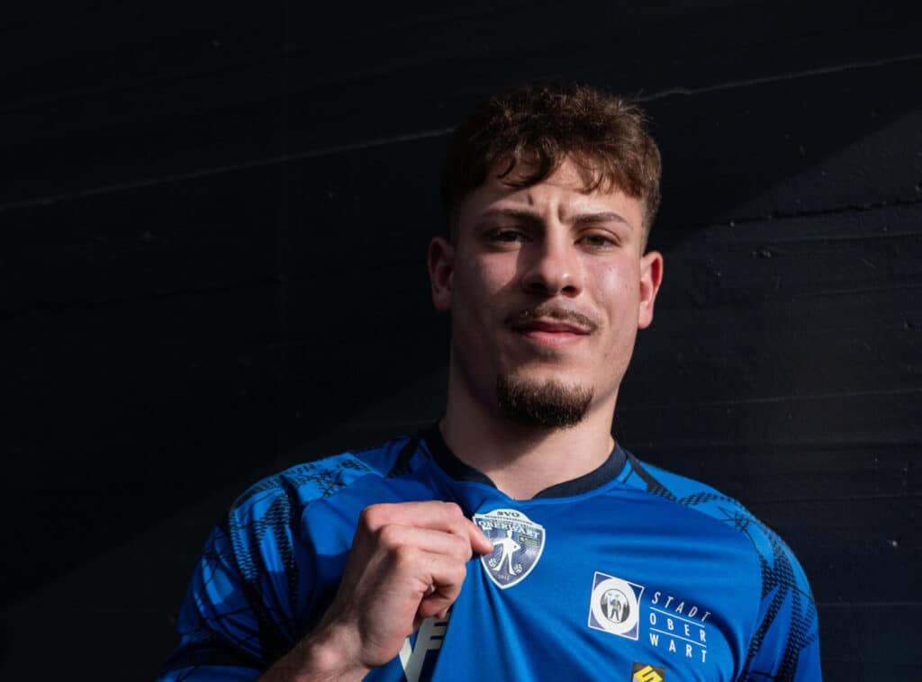 Sportler im blauen Trikot hält Vereinswappen, lächelnd vor dunklem Hintergrund. Teamspirit und Stolz im Vordergrund.