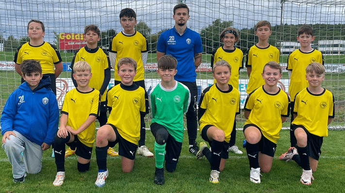 Jugendfußballmannschaft in gelben Trikots posiert auf dem Spielfeld vor einem Tor, Trainer im Hintergrund.