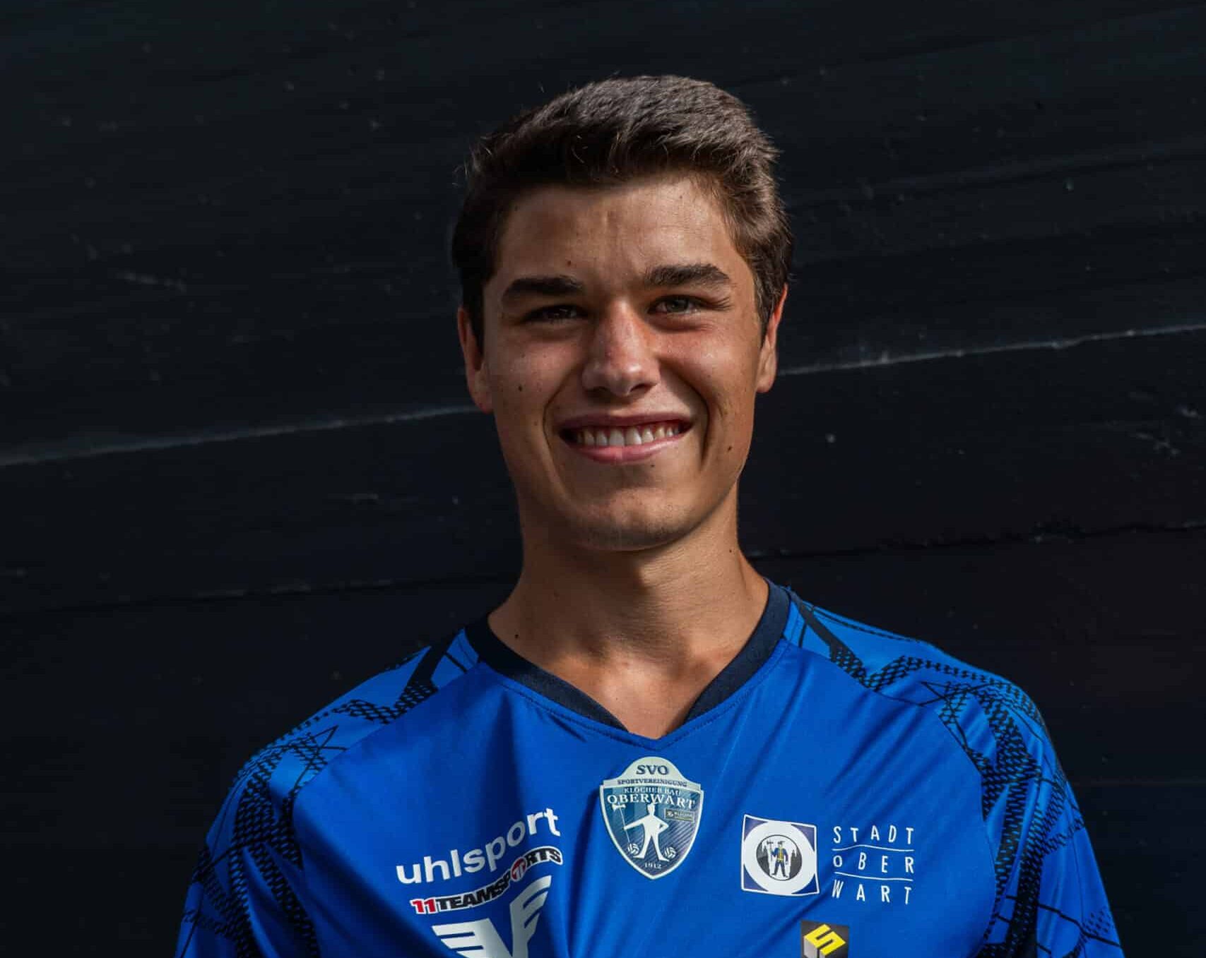 Lächelnder Fußballspieler im blauen Trikot mit Sponsorenlogos, vor dunklem Hintergrund posierend.