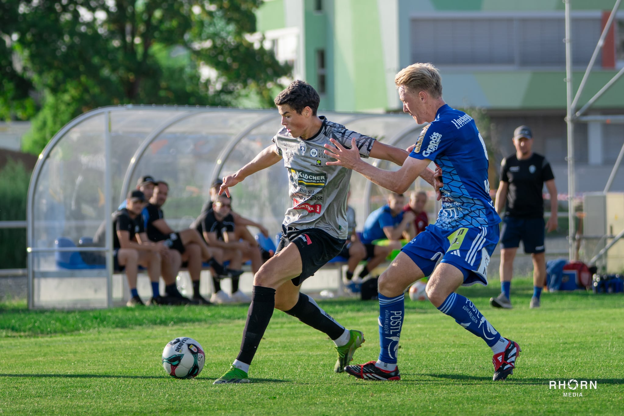 Zwei Fußballspieler im Zweikampf um den Ball während eines Spiels auf grünem Rasen vor Ersatzbank.