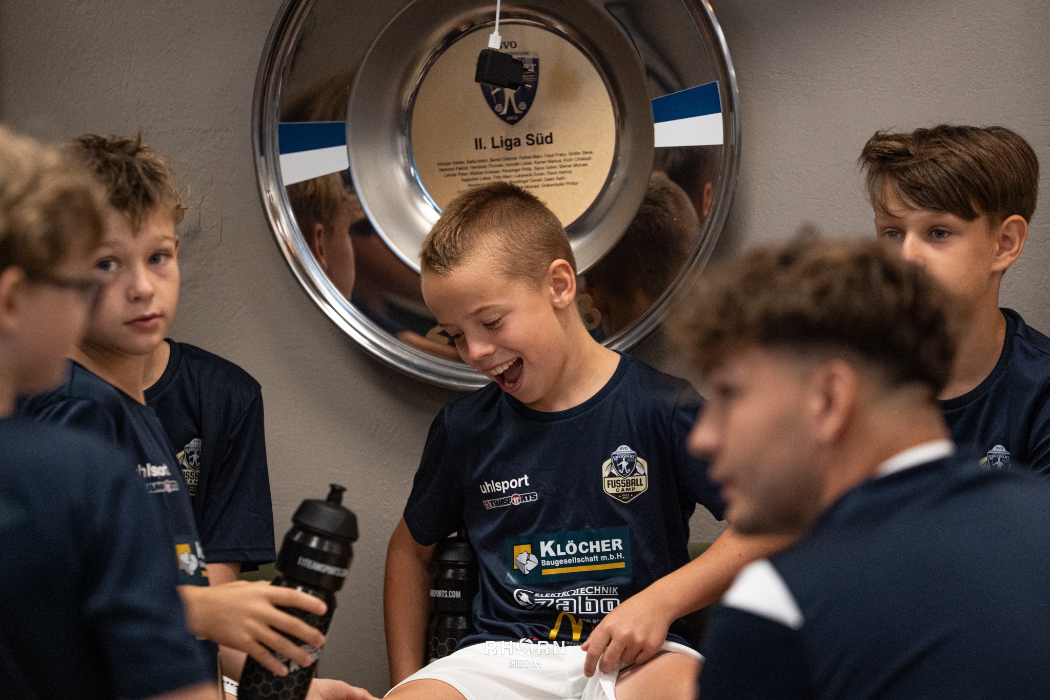 Junge Fußballspieler in der Kabine freuen sich vor einem Spiel, mit einer großen Medaille im Hintergrund.