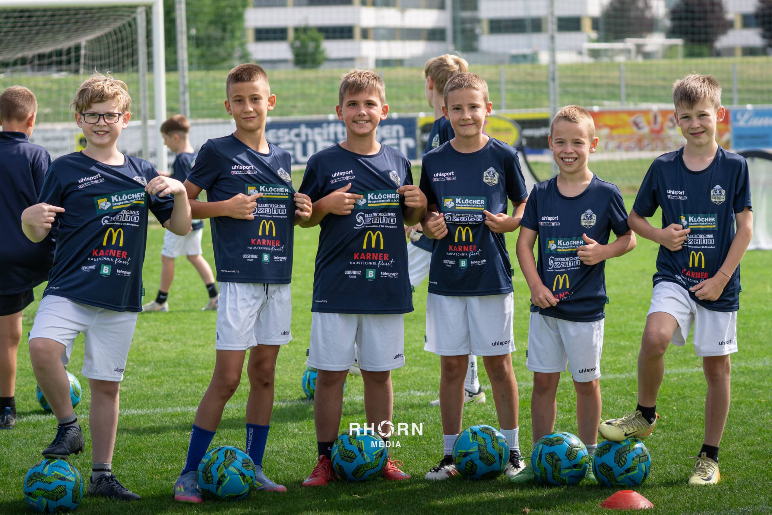 Kinderfußballteam posiert auf dem Spielfeld mit Bällen und Trikots, glücklich auf einer grünen Rasenfläche.
