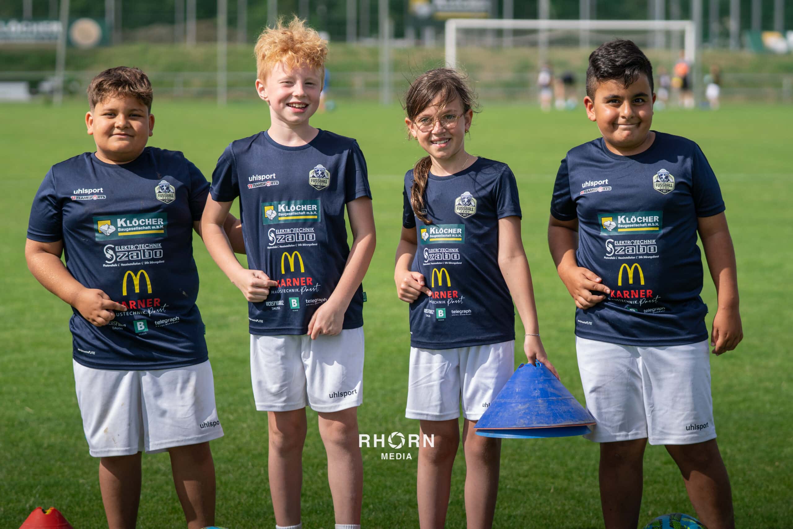 Kinder in Fußballtrikots auf dem Sportplatz lächeln in die Kamera, mit Pylonen in der Hand.