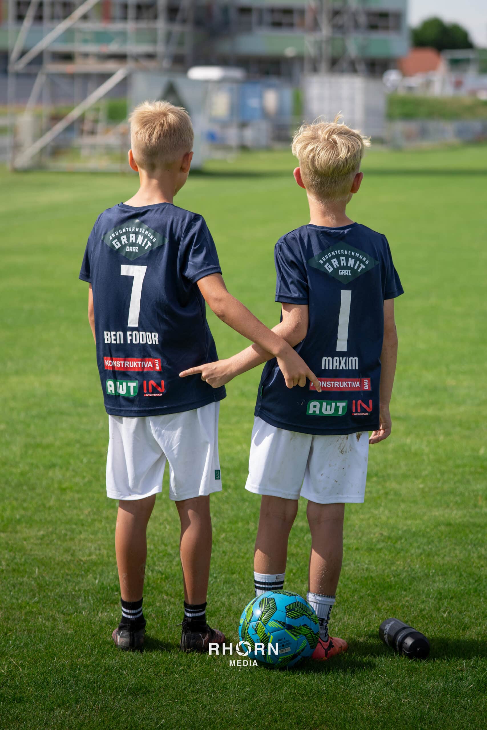 Zwei junge Fußballspieler in blauen Trikots auf dem Rasen, bereit für das Spiel, mit einem bunten Ball.