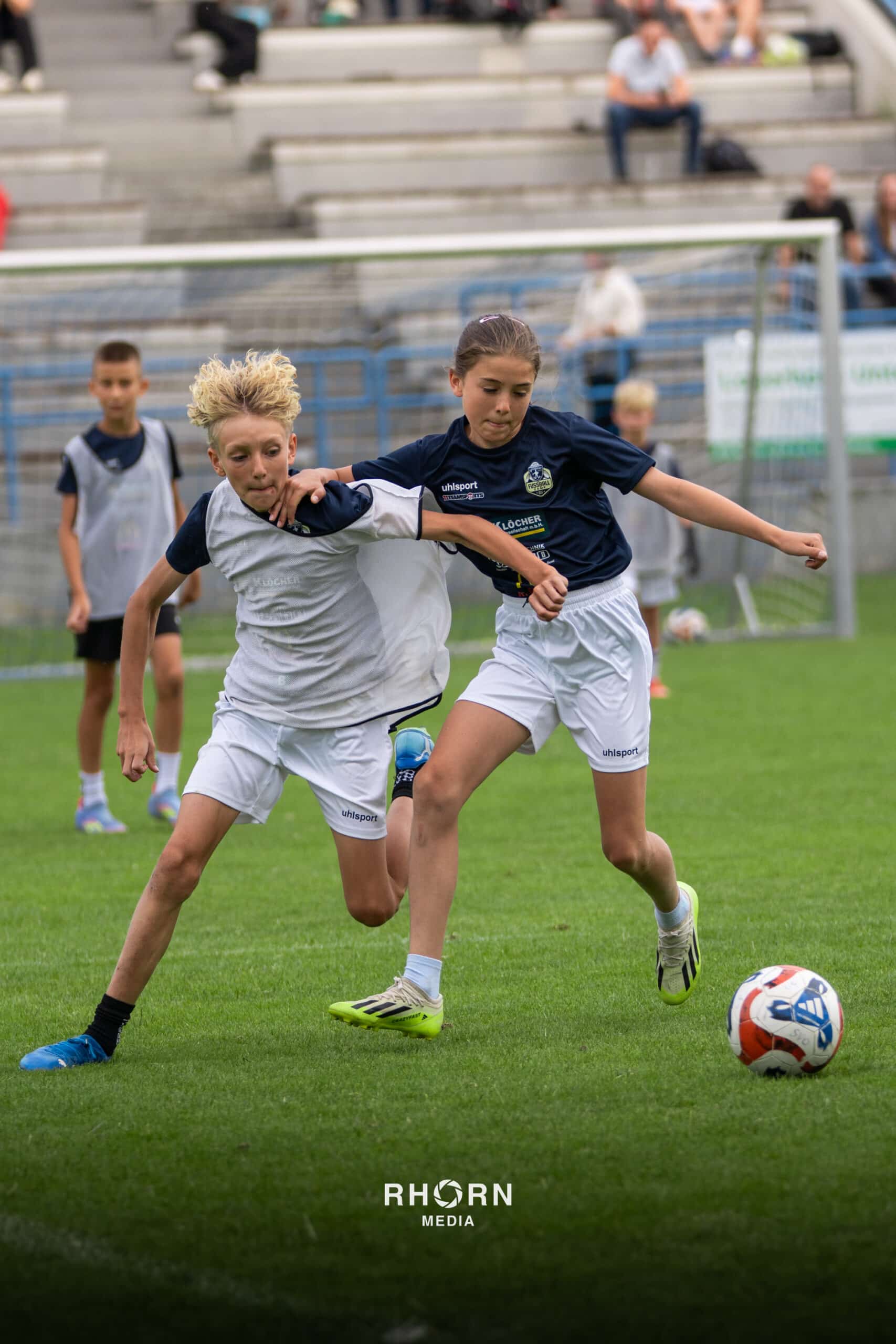 Jugendliche im Fußballspiel auf dem Platz kämpfen um den Ball vor einem Publikum auf der Tribüne.