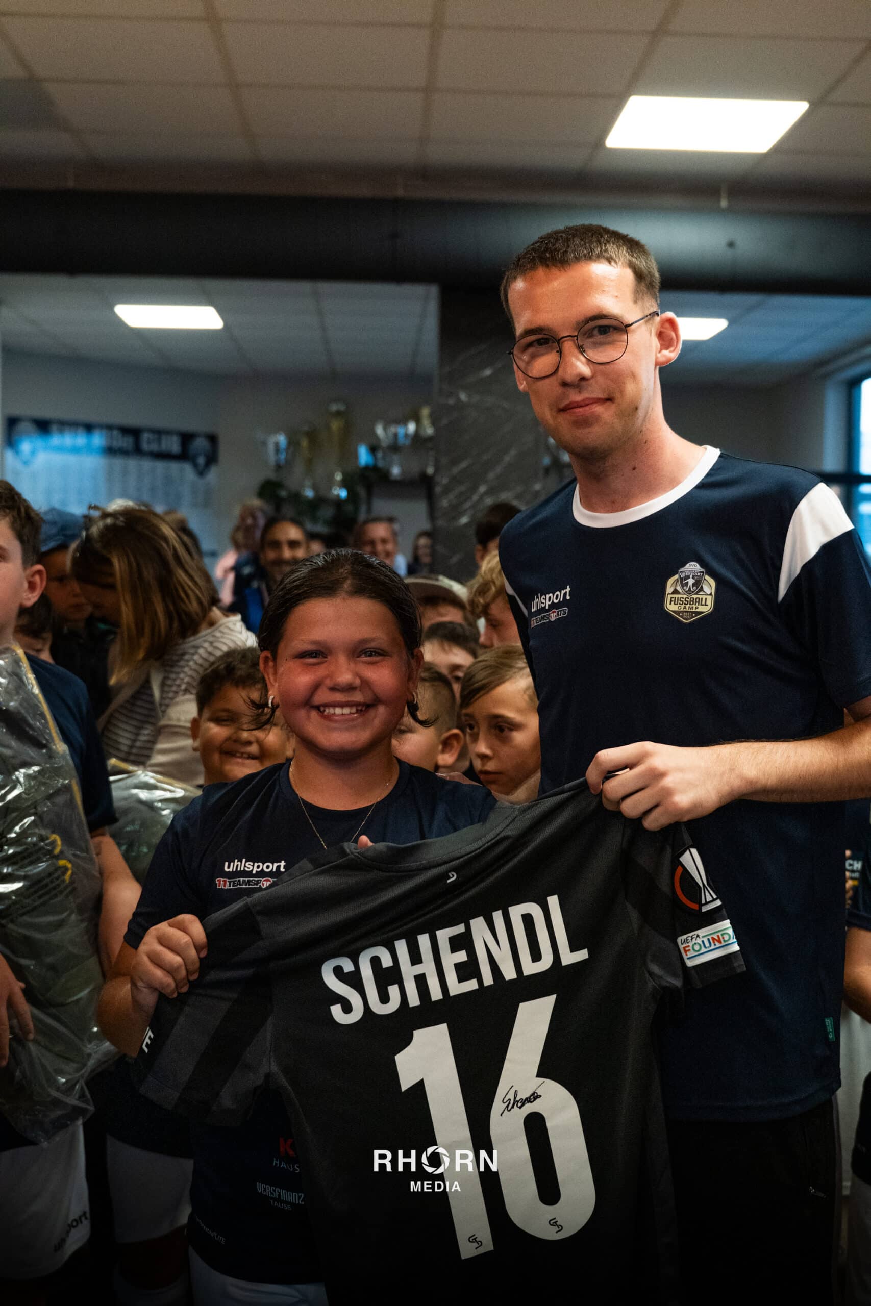 Glückliches Kind hält signiertes Fußballtrikot (Schendl 16) mit Trainer, umgeben von Teamkameraden, im Vereinsraum.