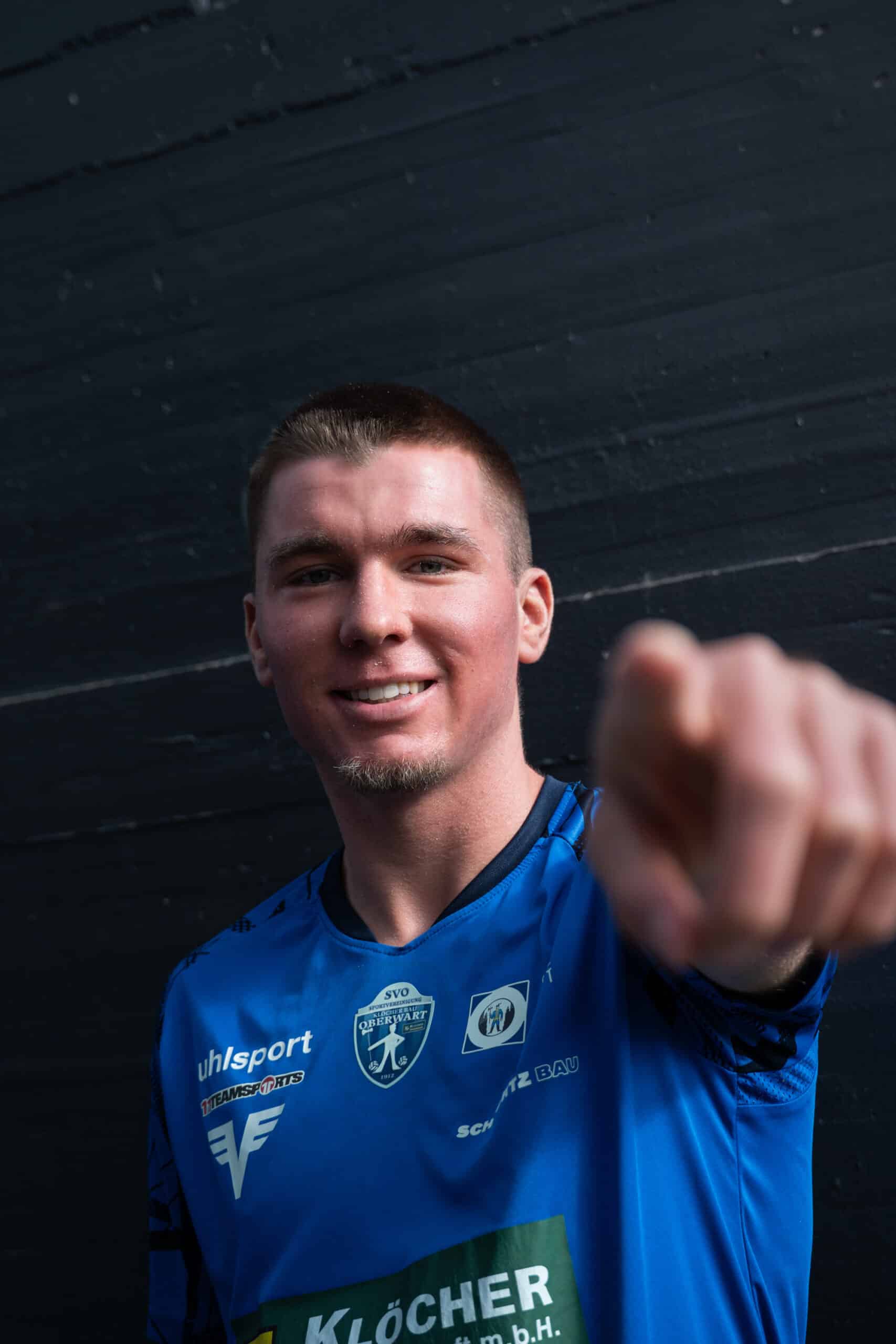 Fußballspieler im blauen Trikot zeigt motivierend in die Kamera vor dunklem Hintergrund.