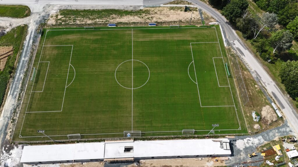 Luftbild eines grünen Fußballfeldes mit sichtbaren Spielfeldmarkierungen und umliegender Landschaft.