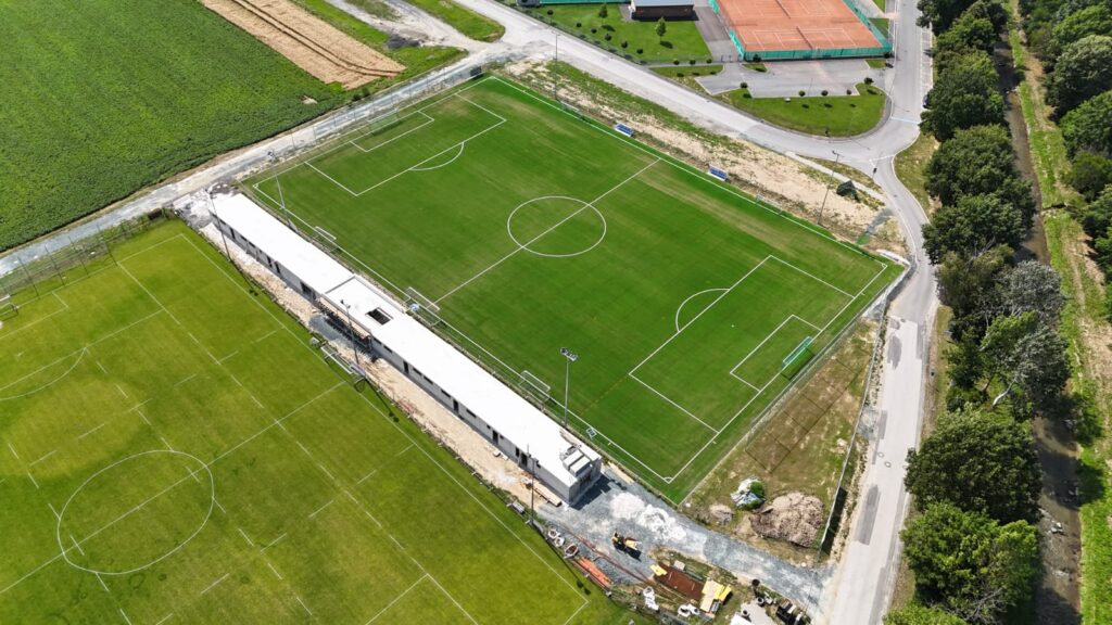 Luftaufnahme eines modernen Sportkomplexes mit Fußballfeld, Tennisplatz und umliegender grüner Landschaft.