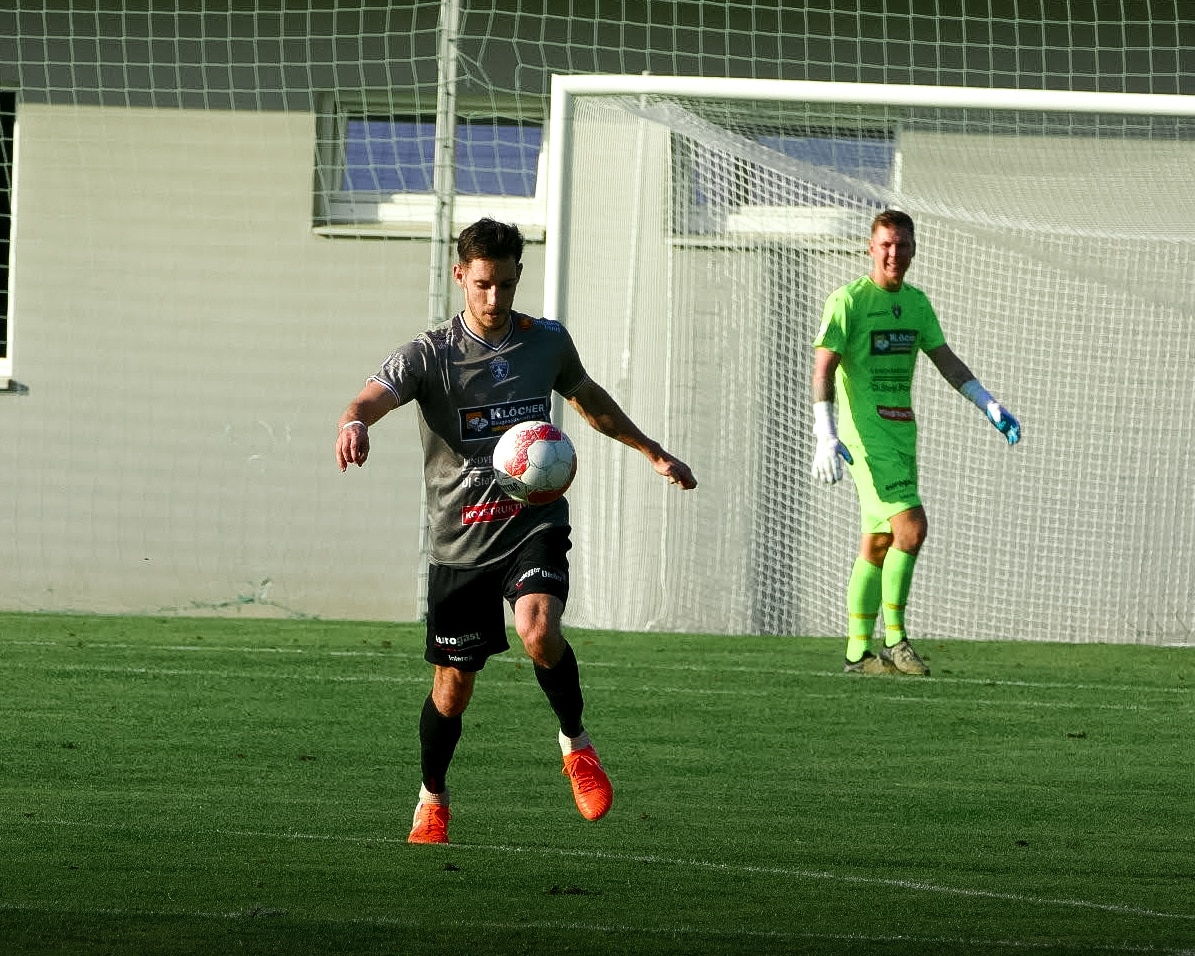 Fußballspieler dribbelt den Ball auf dem Spielfeld, während ein Torhüter im Hintergrund aufmerksam beobachtet.