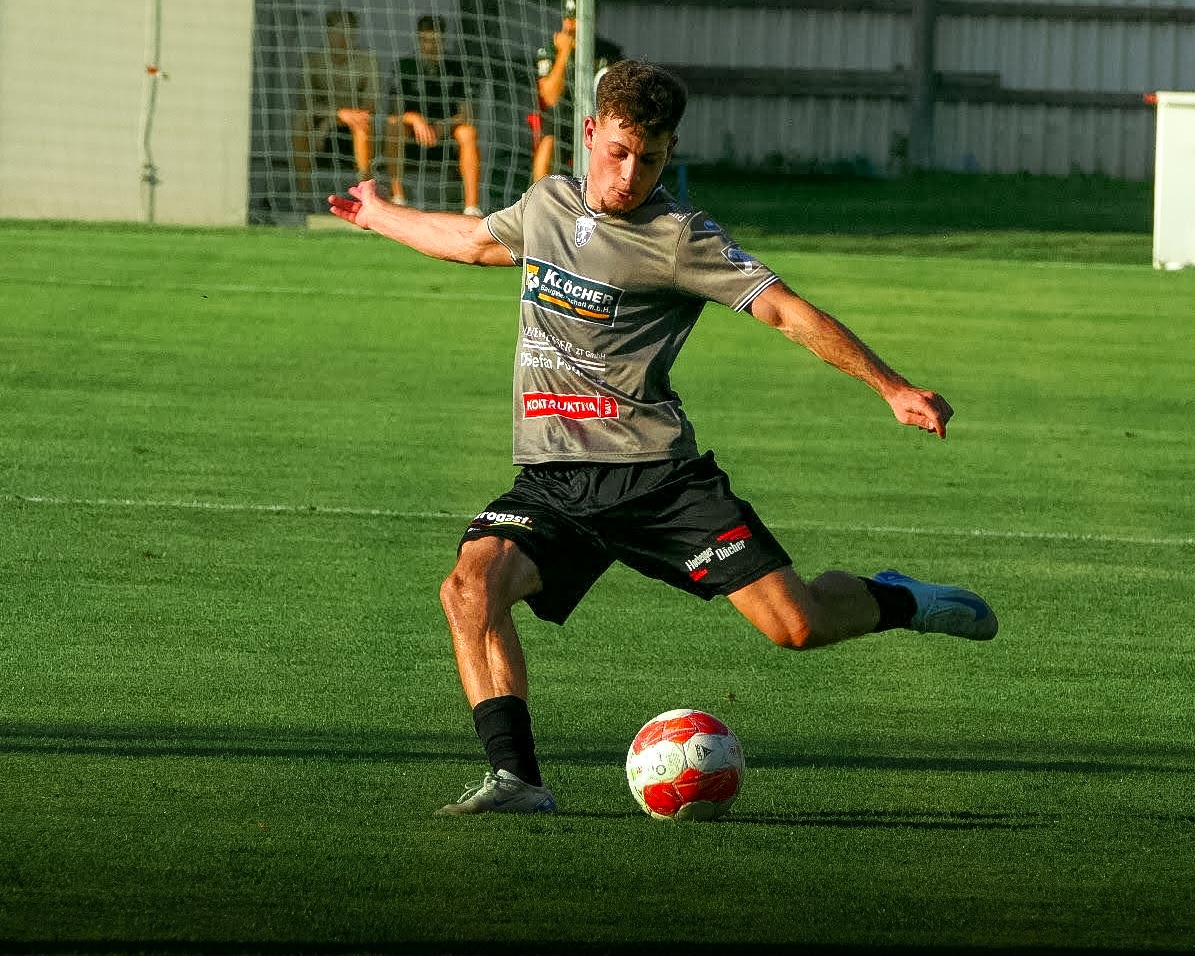 Fußballspieler im grauen Trikot beim Schießen eines Balls auf dem Spielfeld, im Hintergrund ein Tor.