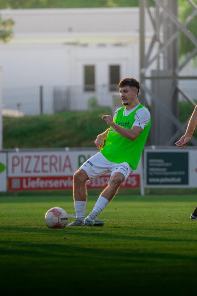 Fußballspieler beim Training auf dem Spielfeld in Aktion mit grünem Trikot und Ball, im Hintergrund Werbebanner.
