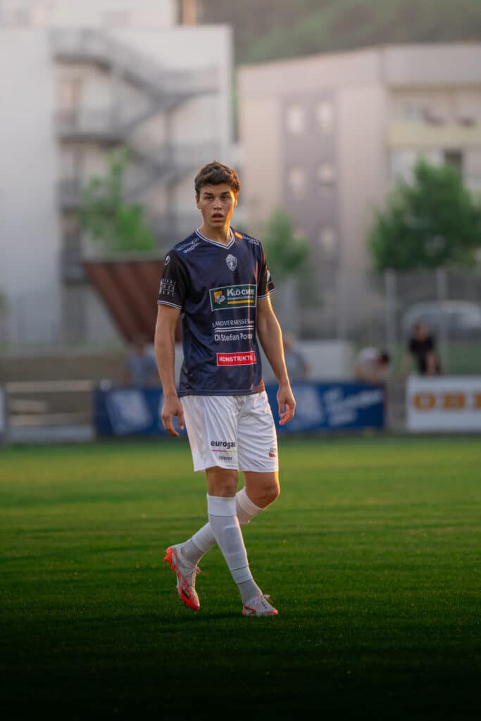 Fußballspieler im blauen Trikot und weißer Hose auf dem Spielfeld vor urbanger Kulisse bei Sonnenuntergang.