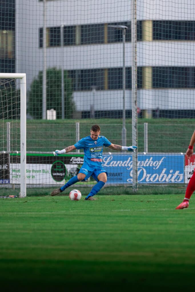 Fußballtorwart im blauen Trikot bereitet sich auf einen Abstoß vom Tor vor, während ein Spieler in Rot zusieht.