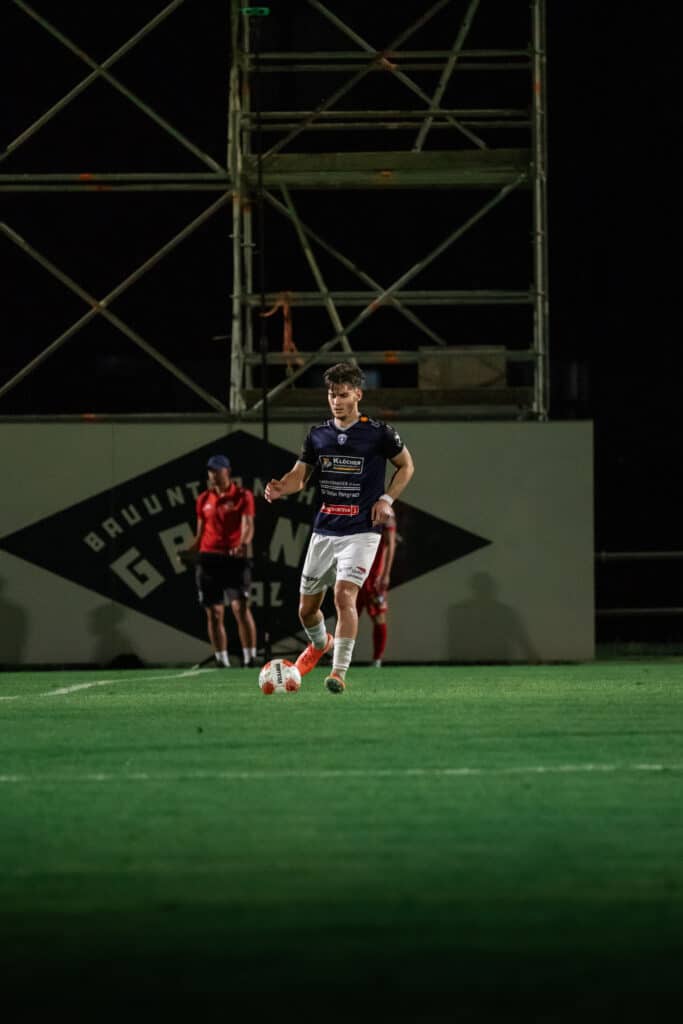 Fußballspieler auf dem Feld bei Nachtspiel, bereit zum Pass auf grünem Rasen mit Flutlicht im Hintergrund.