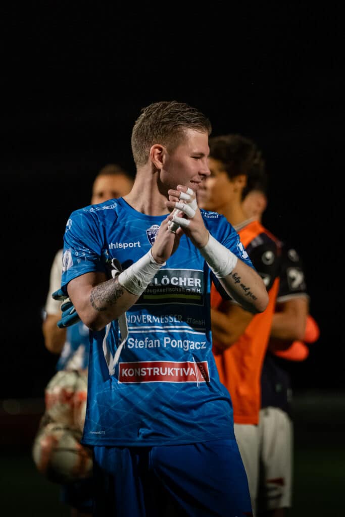 Fußballspieler in blauem Trikot klatscht mit Team im Hintergrund bei Nachtspiel, emotionale Teamunterstützung.