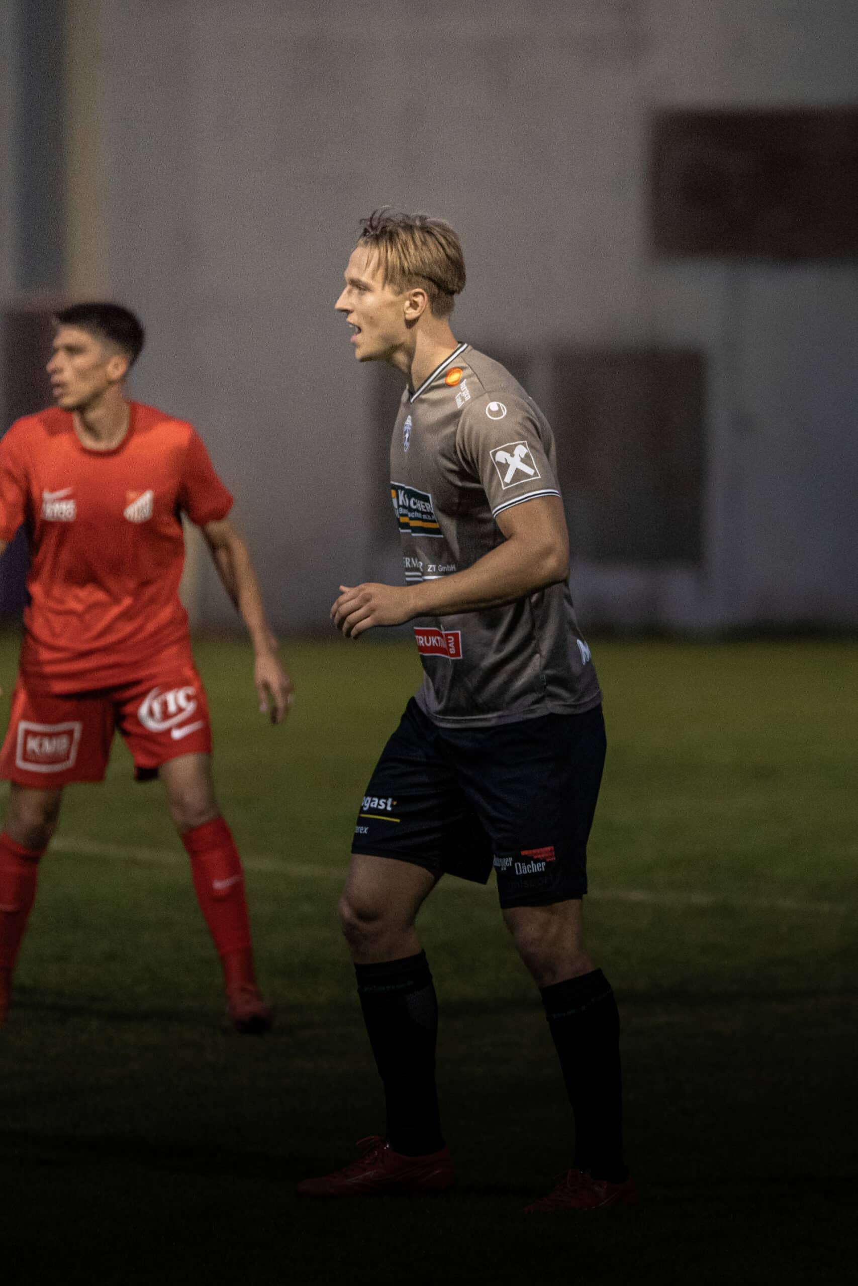 Fußballspieler im grauen Trikot im Spiel, konzentriert auf den Ball, mit Gegner in rotem Trikot beim Nachtspiel.
