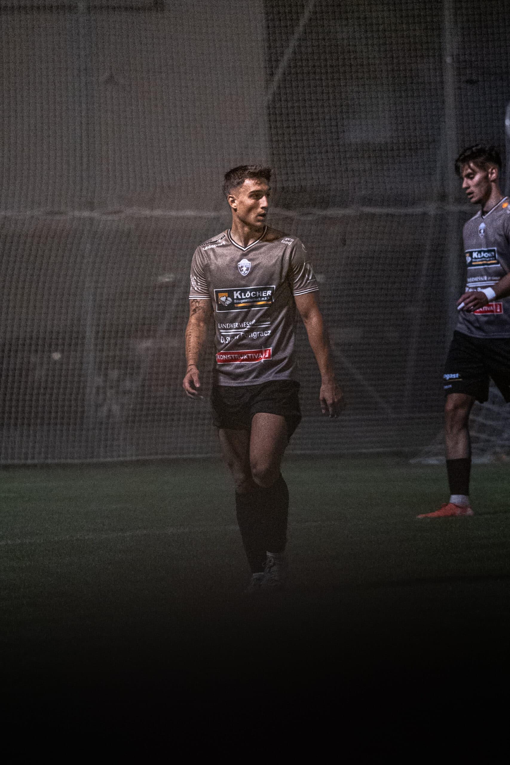 Fußballspieler bei Nacht im Spiel, tragen graues Trikot auf dem Spielfeld, konzentriert und bereit für die Aktion.