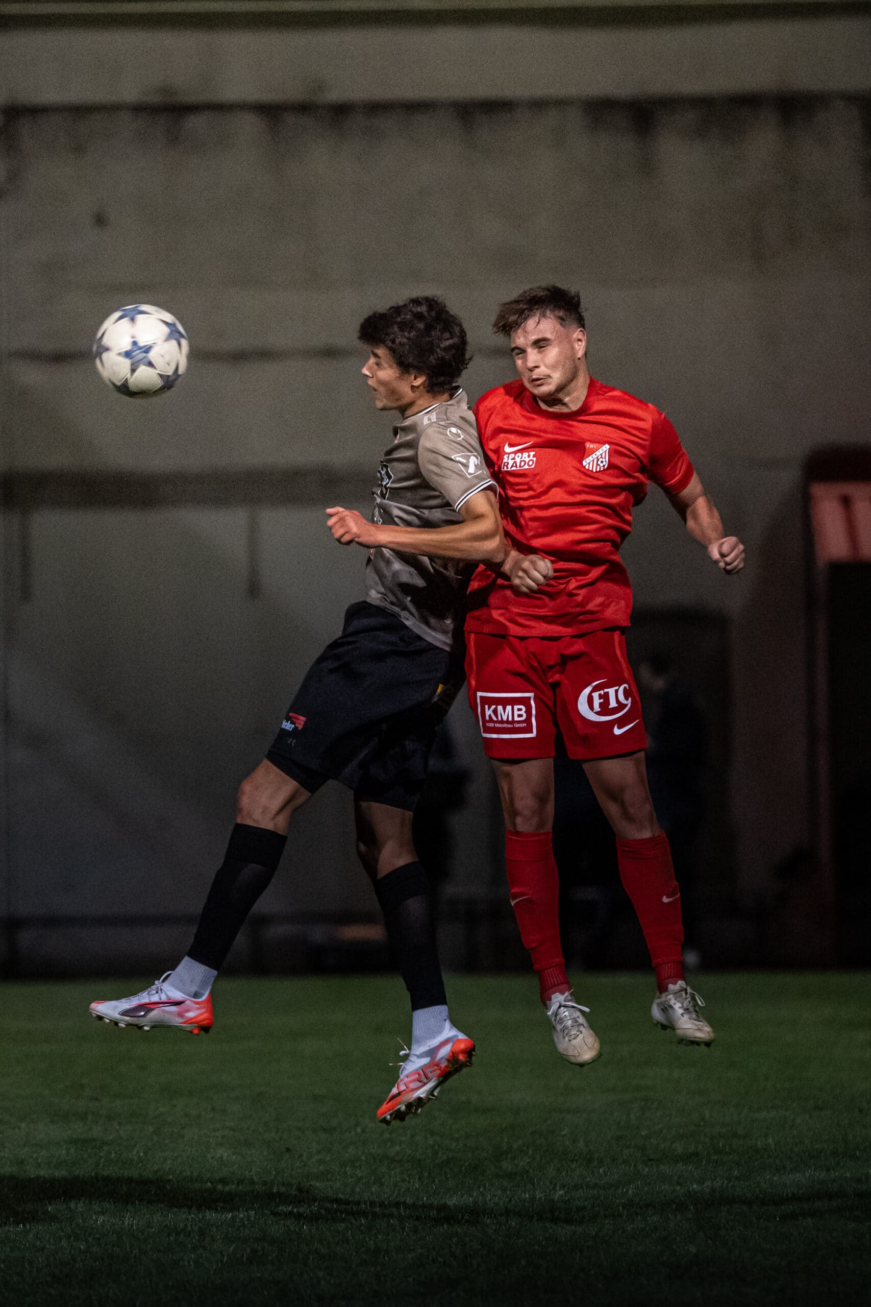 Zwei Fußballspieler im Luftduell um den Ball auf einem nächtlichen Spielfeld. Wettbewerbsintensität und Athletik.
