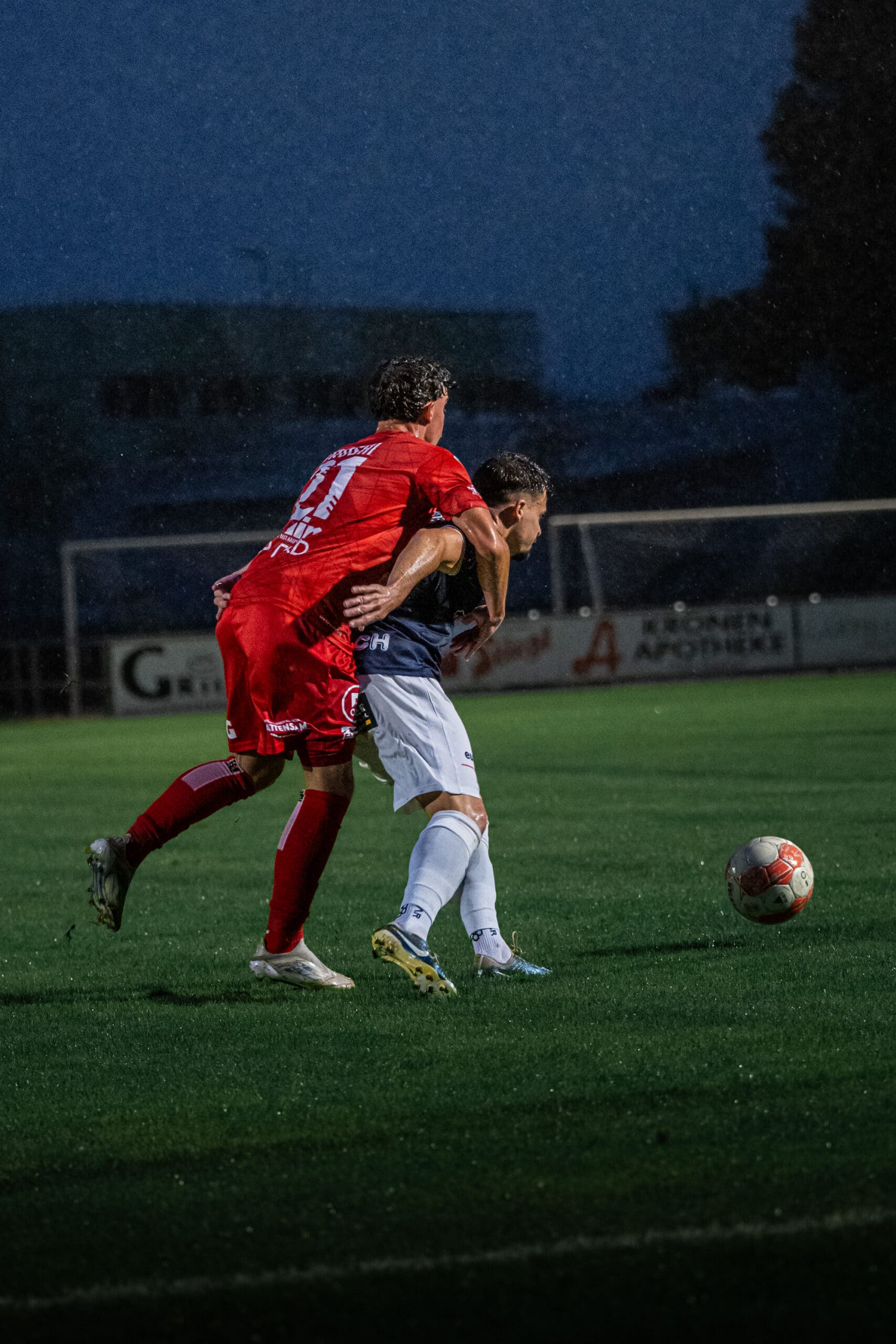 Zwei Fußballspieler kämpfen im Regen um den Ball auf einem nassen Spielfeld, intensive Wettkampfatmosphäre.
