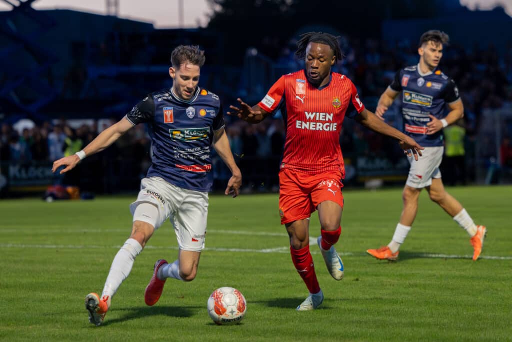 Zwei Fußballspieler im Duell während eines Spiels, einer in blau-weiß, der andere in rot, verfolgen den Ball.