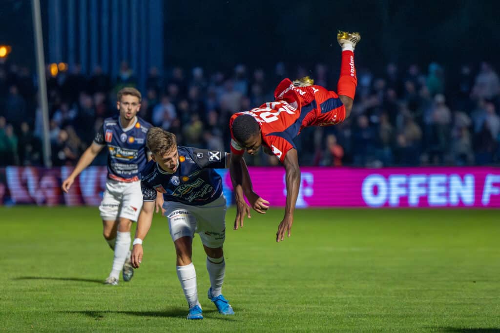 Fußballspiel, Spieler in Rot macht artistische Bewegung, während er von Gegner in Blau herausgefordert wird.