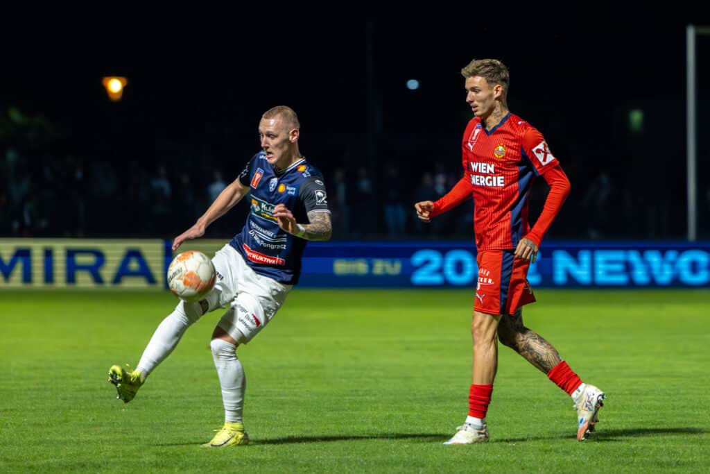 Fußballspiel bei Nacht: Spieler in blauem und rotem Trikot im Zweikampf um den Ball auf dem Spielfeld.