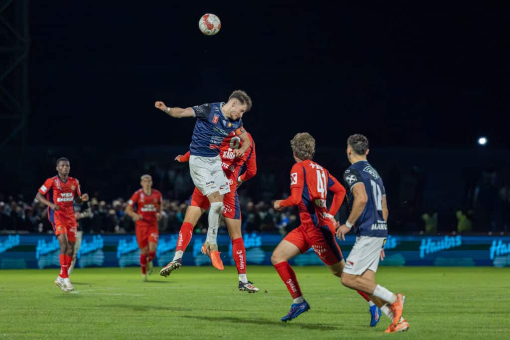 Fußballspieler im Lufteinsatz während eines Nachtspiels, umgeben von Gegnern in roter Kleidung.