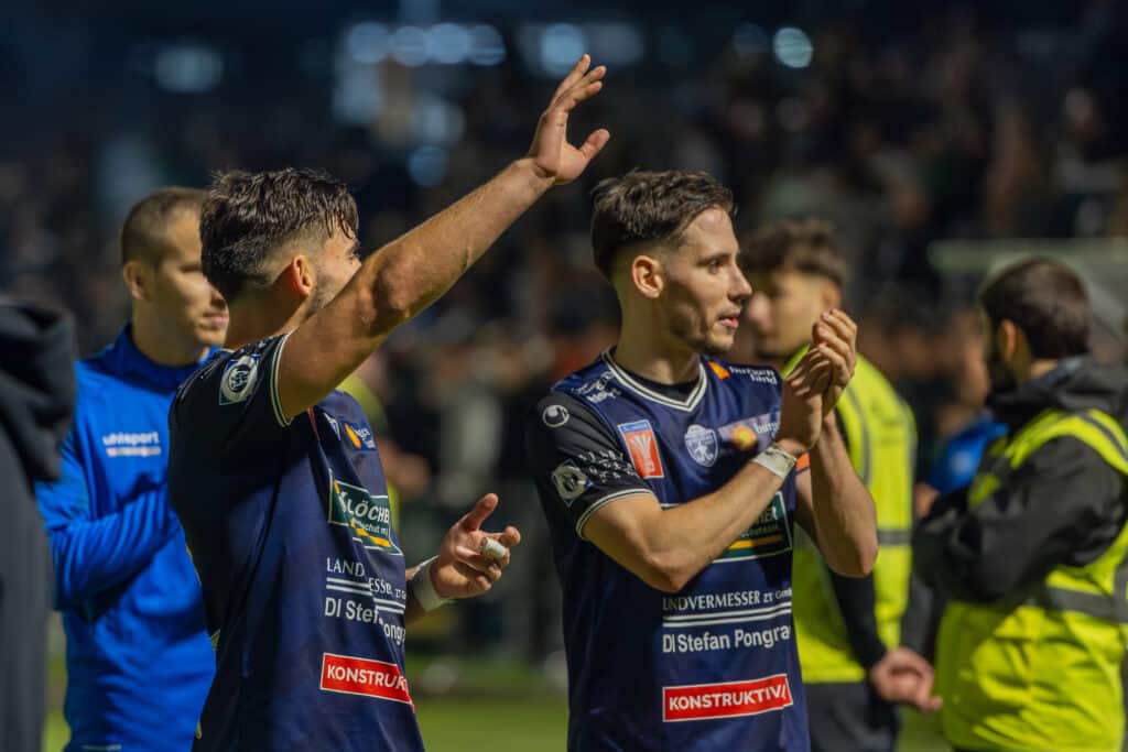 Fußballspieler feiern nach einem Spiel auf dem Feld, klatschend und jubelnd mit den Fans.