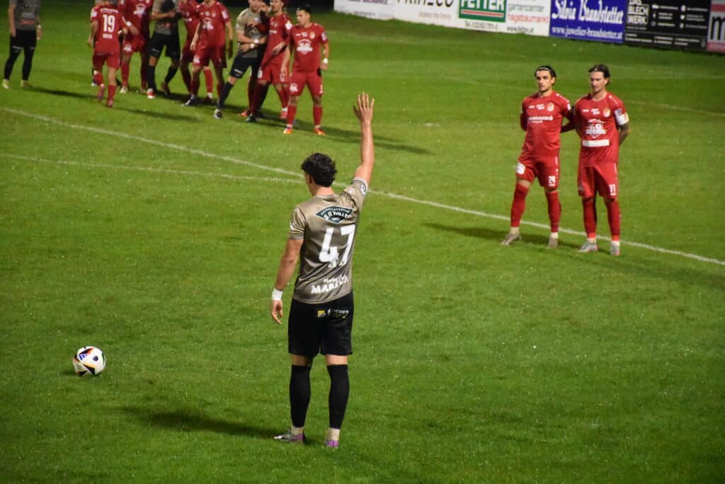 Fußballspieler bereitet Freistoß vor, Gegenspieler stellen Mauer auf, Nachtspiel auf grünem Rasen im Stadion.