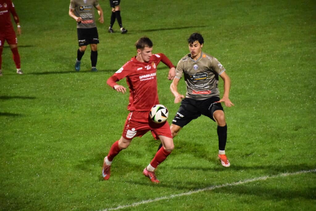 Zwei Fußballspieler kämpfen auf regennassem Rasen in einem Match um den Ball, konzentriert und bereit für die nächste Aktion.