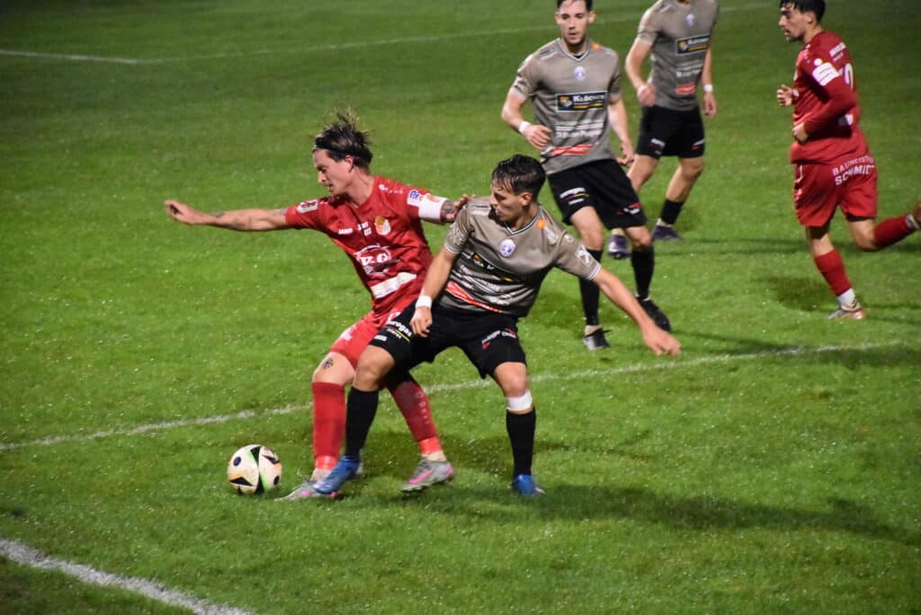 Zwei Fußballspieler kämpfen auf regennassem Rasen um den Ball. Im Hintergrund weitere Spieler in Aktion.
