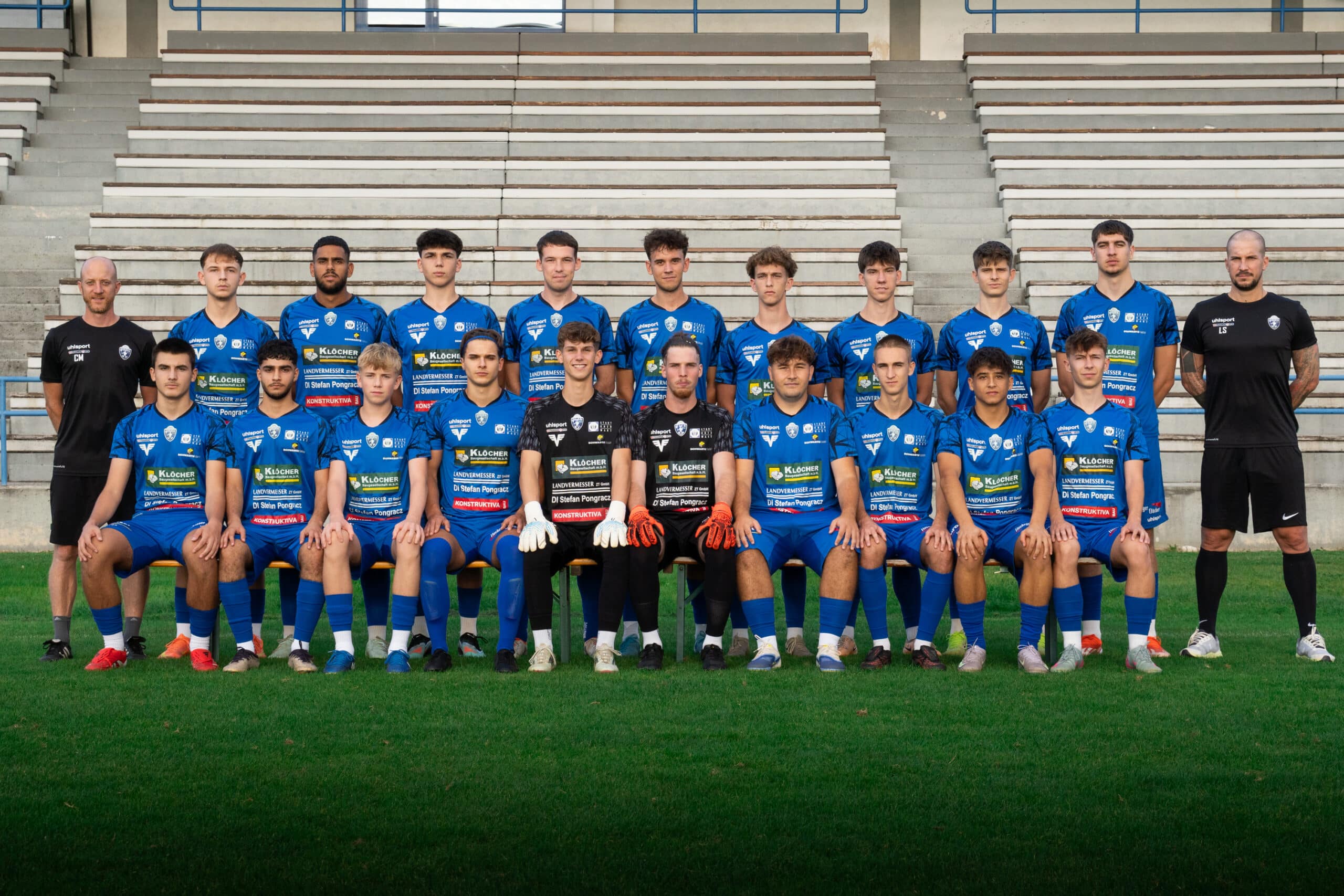 Fußballmannschaft in blauen Trikots, Gruppenfoto im Stadion auf Rasen mit Trainern links und rechts stehend.