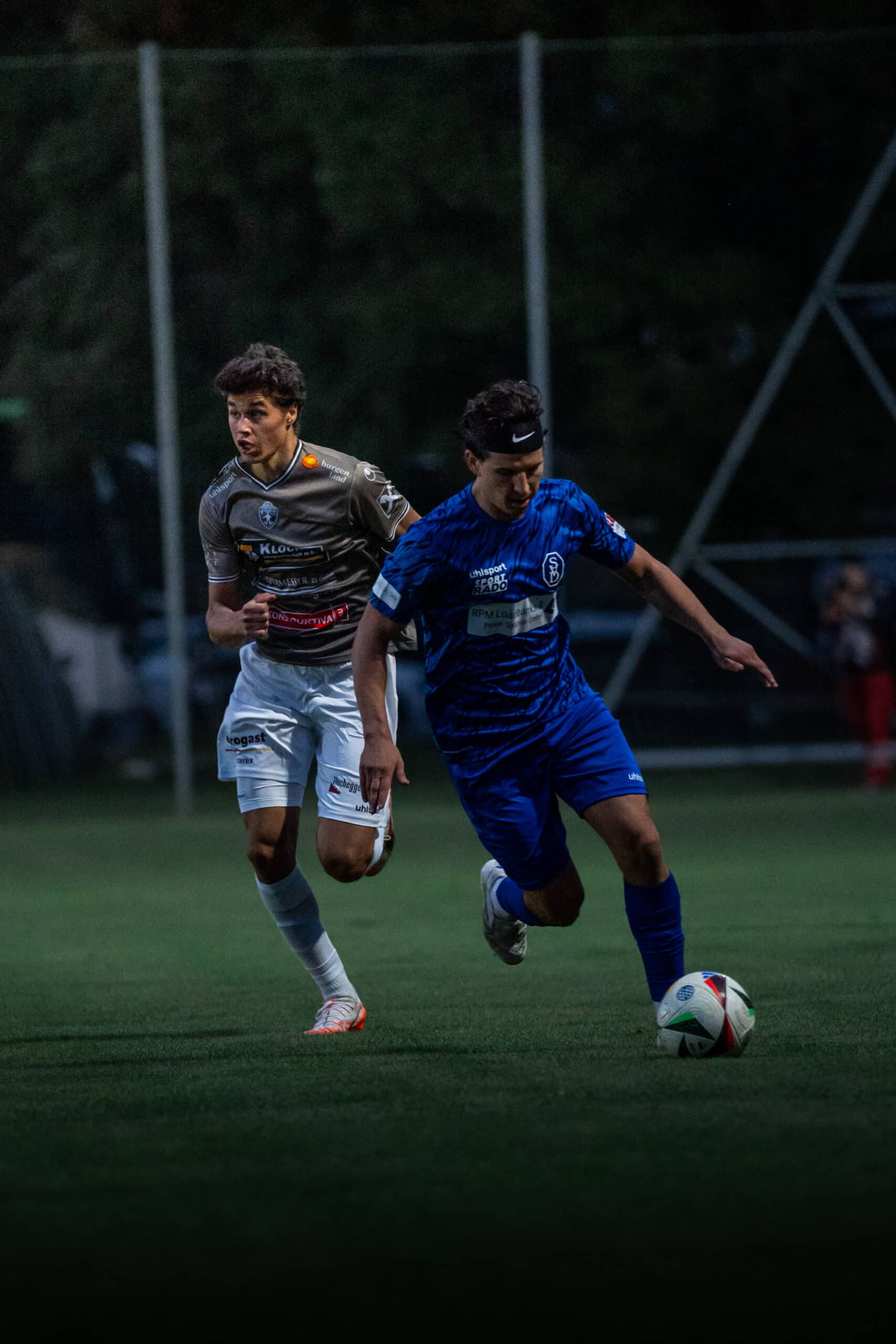 Zwei Fußballspieler im Wettkampf auf dem Spielfeld bei Nacht. Spieler in blauer und grauer Uniform sprinten um den Ball.