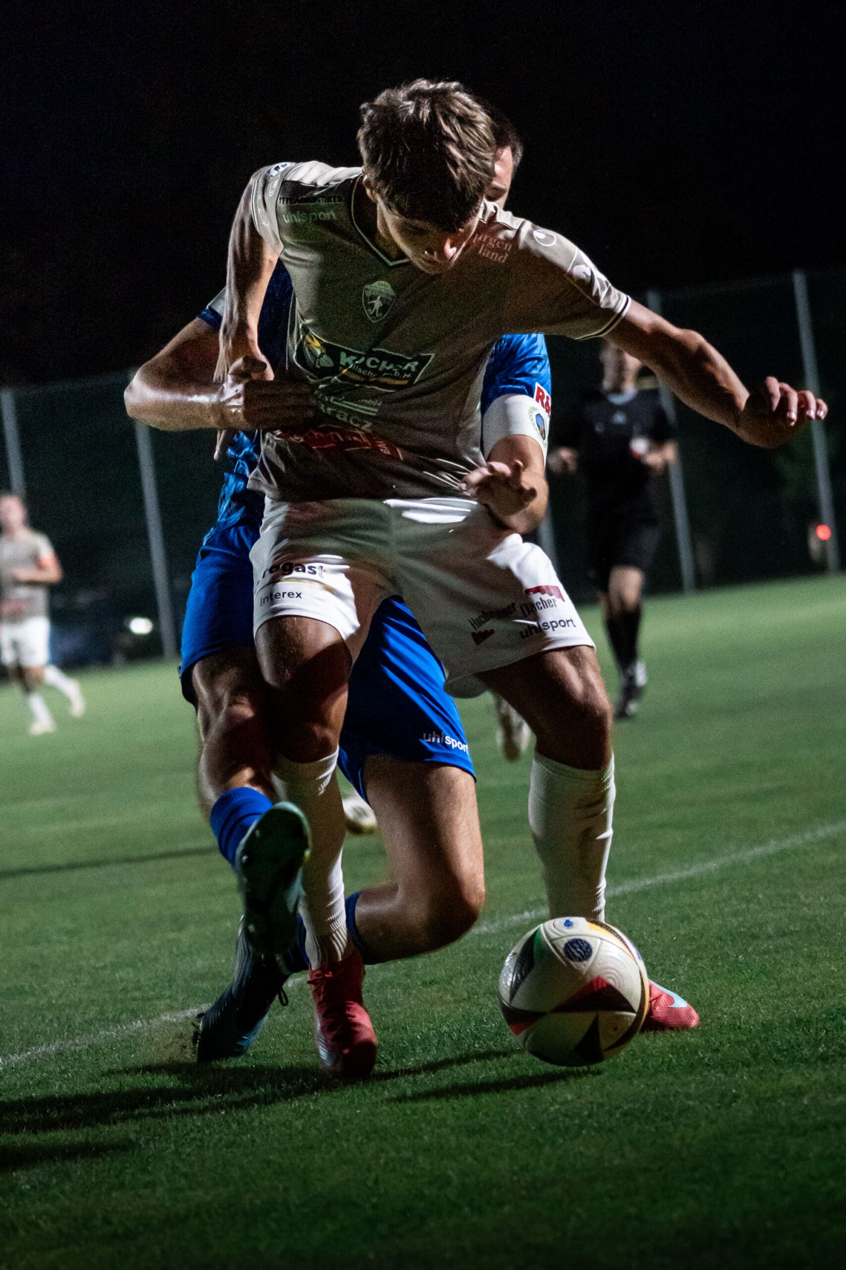 Zwei Fußballspieler kämpfen bei einem nächtlichen Spiel um den Ball auf einem beleuchteten Spielfeld.