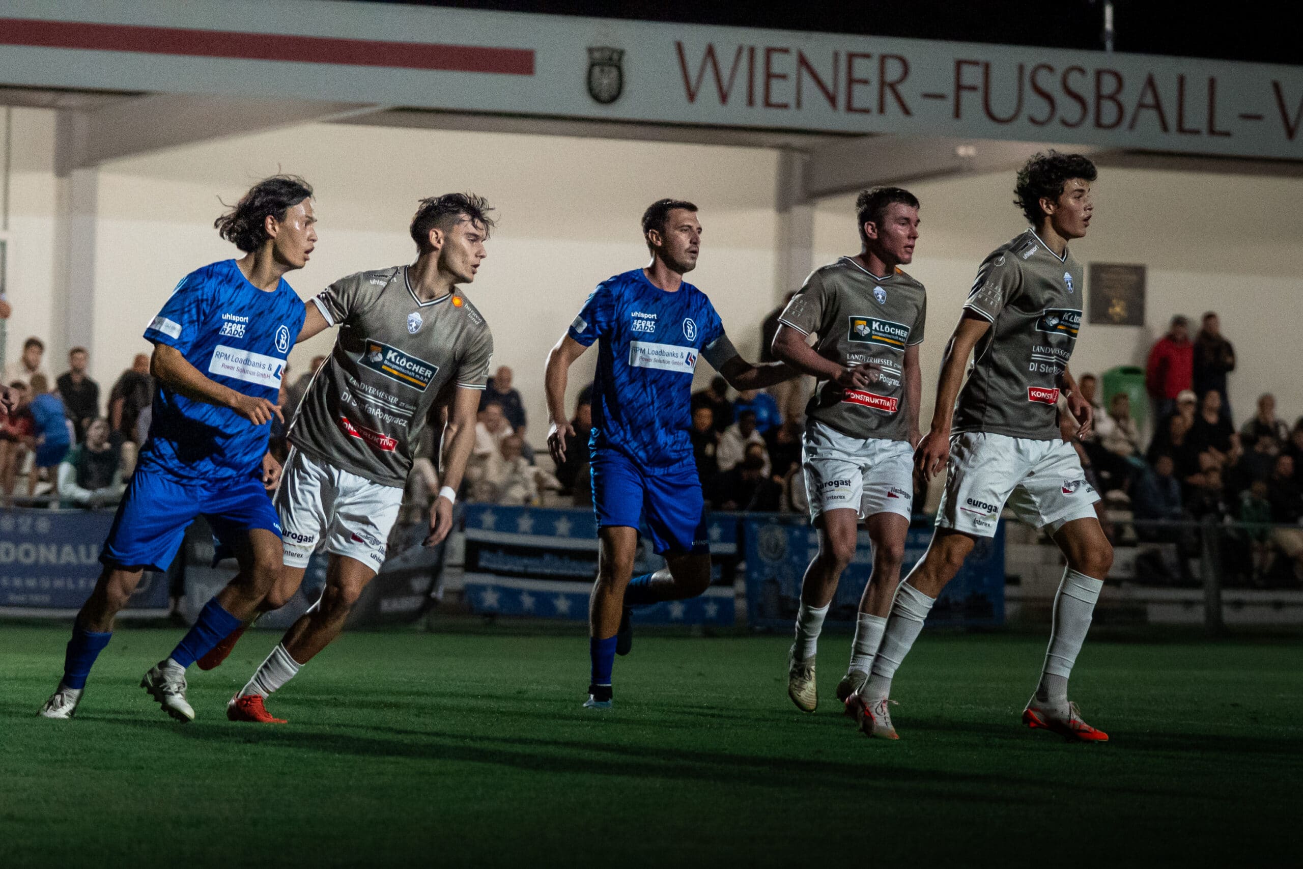 Fußballspiel bei Nacht im Wiener Stadion mit Spielern in blauen und grauen Trikots in intensiver Aktion.