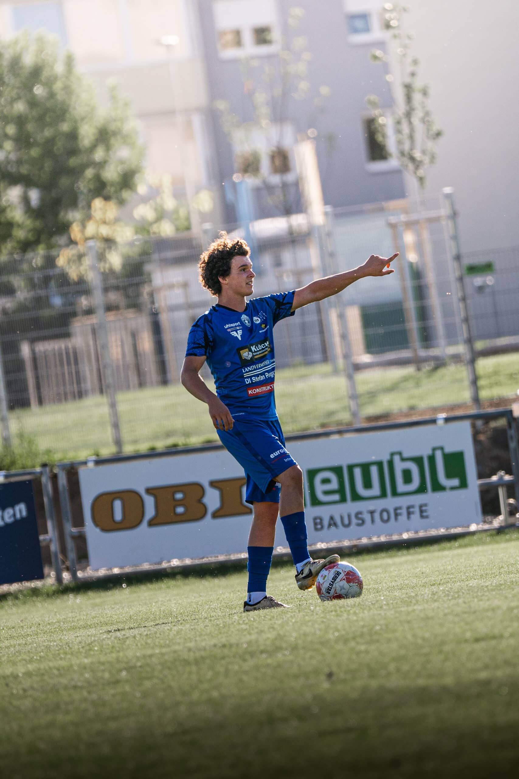 Fußballspieler in blauem Trikot dirigiert während eines Spiels auf dem Feld, mit Fokus auf Ballkontrolle und Teamkoordination.