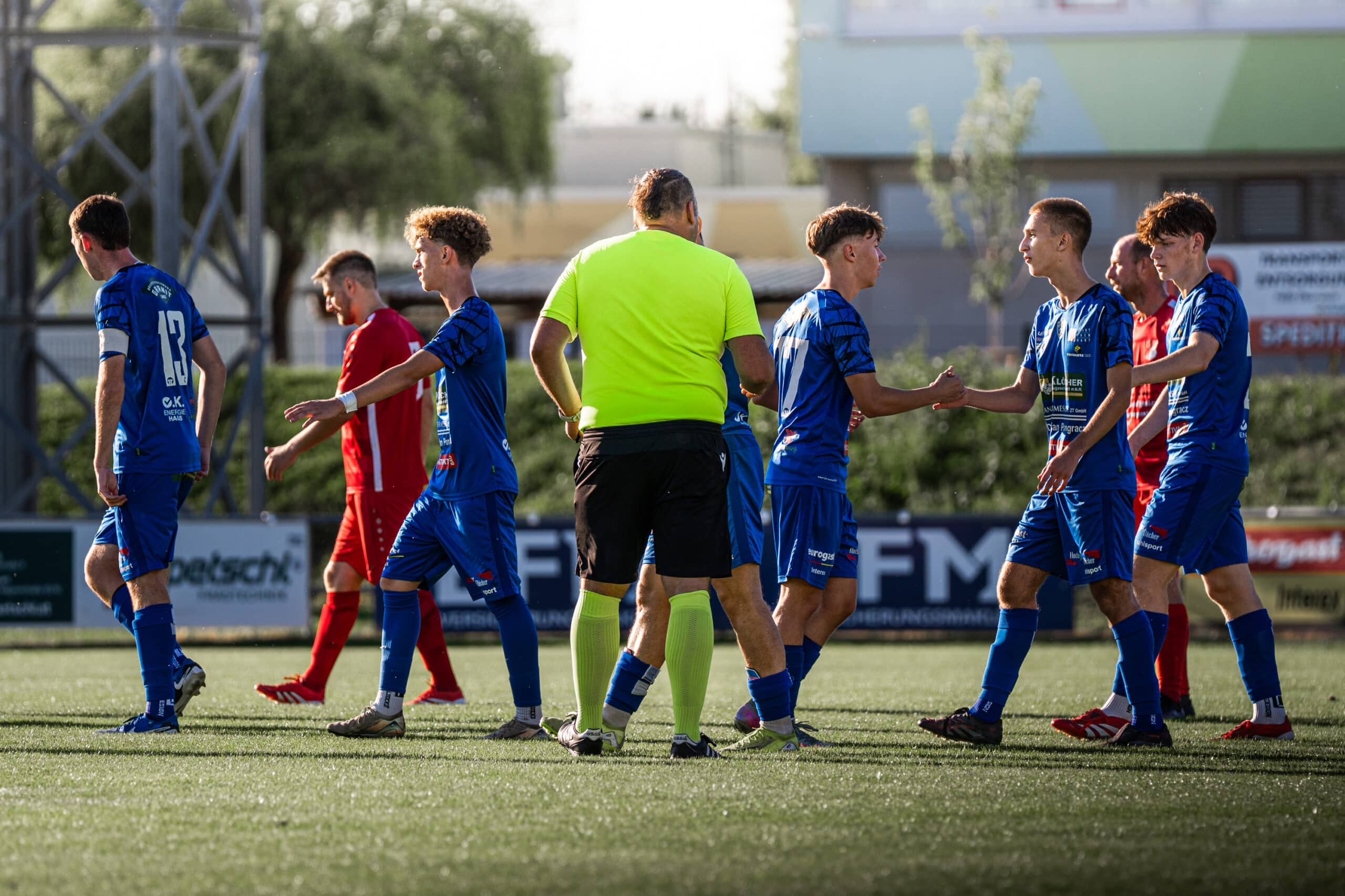 Fußballspieler und Schiedsrichter auf dem Spielfeld beim Handschlag nach dem Spiel bei sonnigem Wetter.