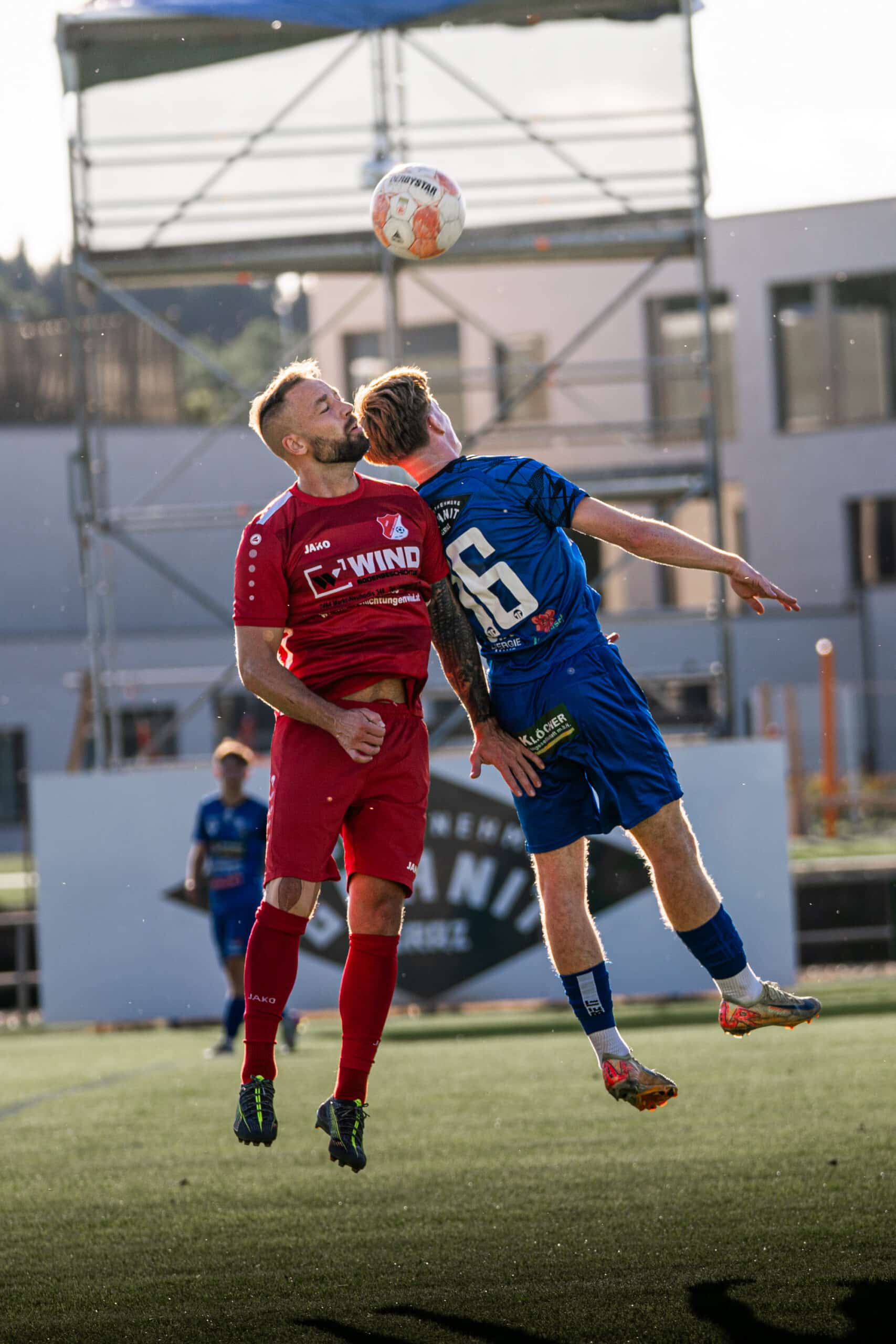 Zwei Fußballspieler im Luftzweikampf um den Ball während eines Spiels auf einem Sportplatz.