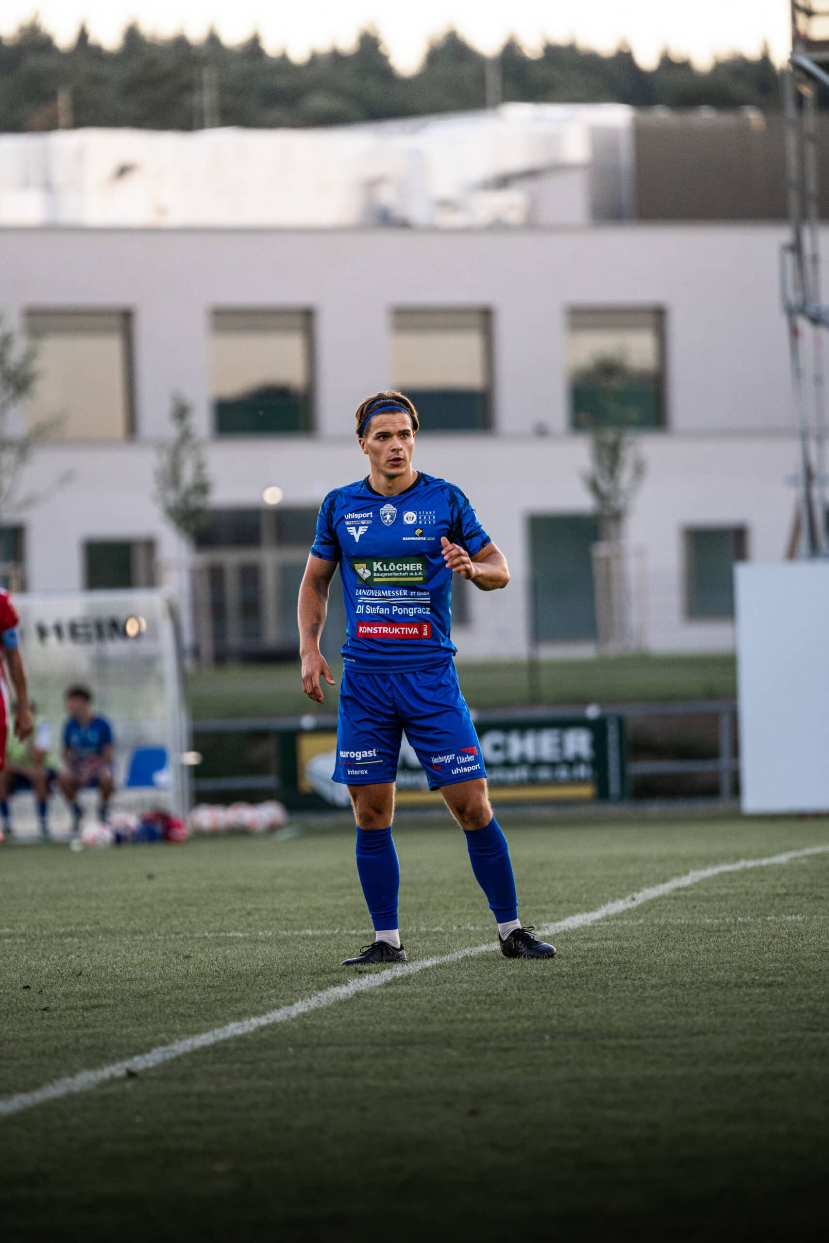 Fußballspieler in blauem Trikot auf dem Spielfeld, Gebäude im Hintergrund.