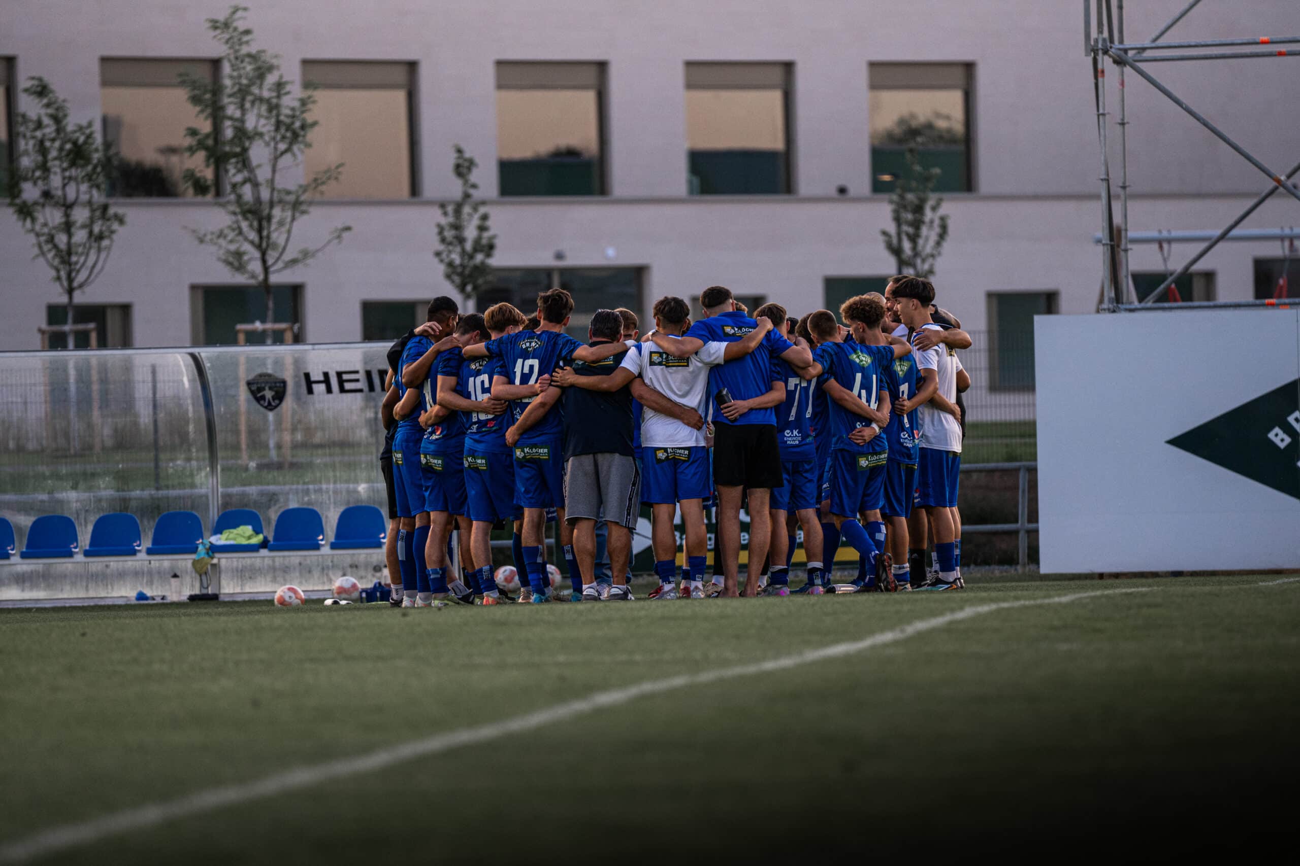Fußballteam in blauen Trikots versammelt sich für Motivationskreis auf dem Spielfeld vor einem Spiel.