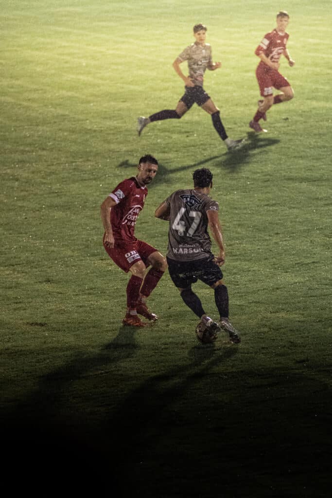 Fußballspiel bei Nacht: Zwei Spieler kämpfen um den Ball auf nassem Rasen, einsatzbereit und konzentriert.