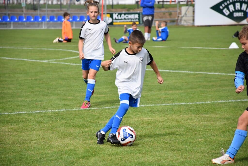 Kinder spielen Fußball auf einem grünen Spielfeld bei sonnigem Wetter, tragen weiße Trikots und blaue Shorts.