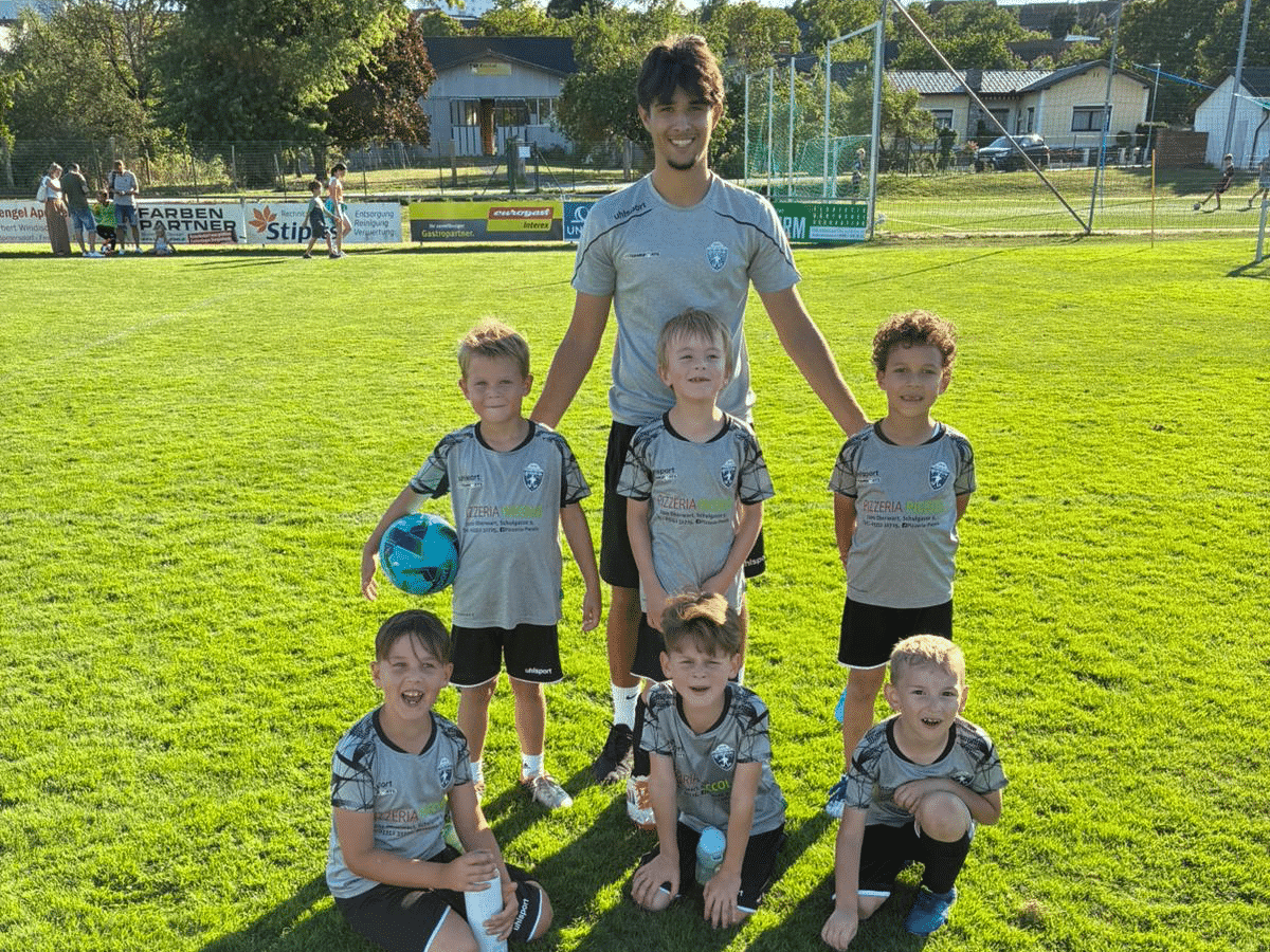 Kinderfußballteam posiert auf dem Sportplatz an einem sonnigen Tag. Trainer steht hinter sechs Spielern.