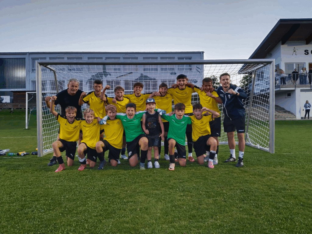 Jugendfußballteam jubelt vor einem Tor auf dem Spielfeld, Trainer und Spieler in gelben Trikots lächeln.