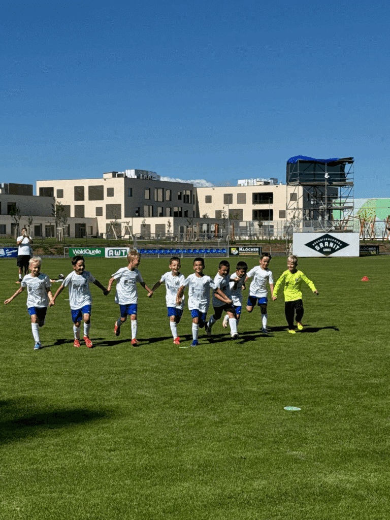 Kinder-Fußballteam läuft Hand in Hand auf einem Sportplatz bei sonnigem Wetter.