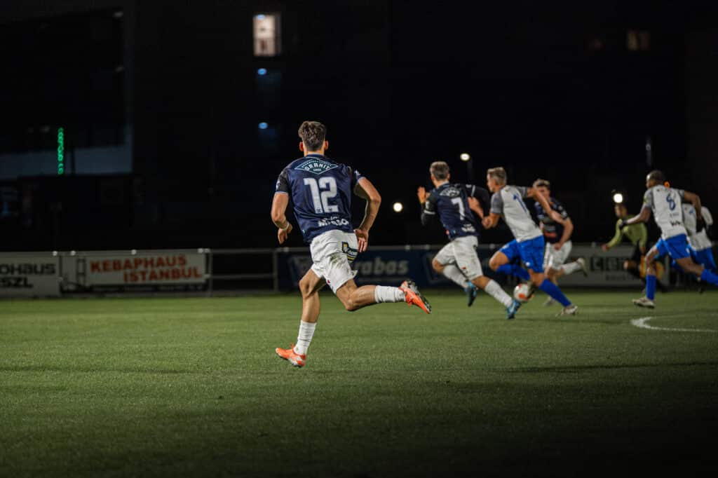 Fußballspiel bei Nacht: Spieler in blauen Trikots sprinten auf dem Feld, dynamische Actionaufnahme.