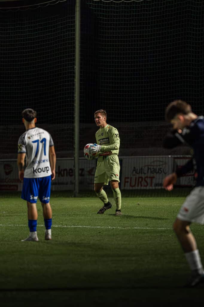 Ein Torwart hält den Ball, während Spieler auf einem nächtlichen Fußballfeld agieren.