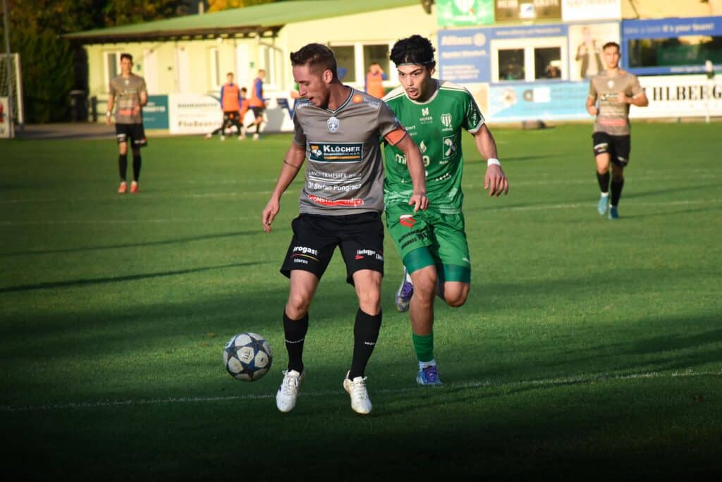 Zwei Fußballspieler im Zweikampf um den Ball auf einem sonnigen Spielfeld während eines Matches, weitere Spieler im Hintergrund.